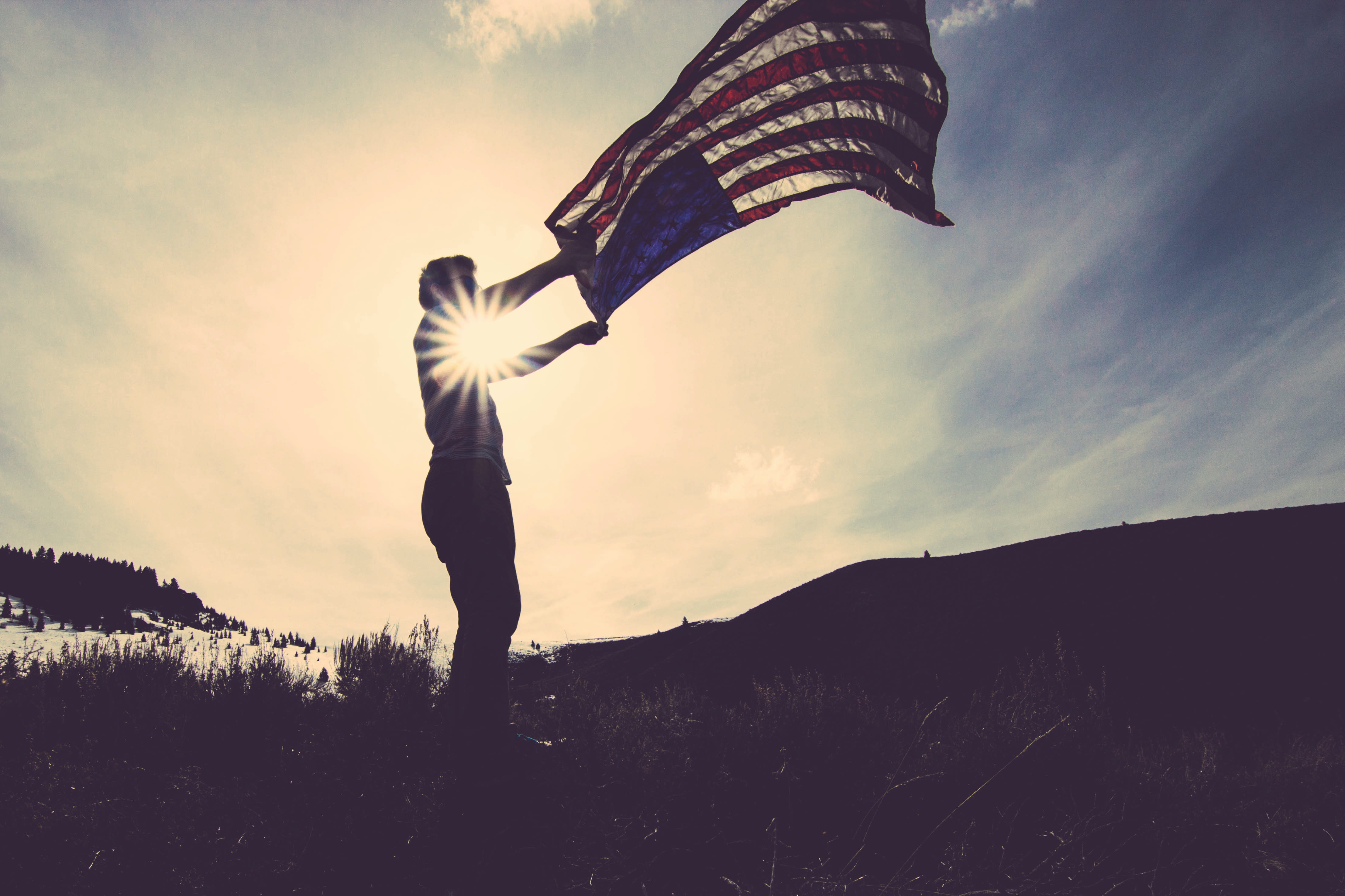 A picture of a man holding an american flag up, with the sun in the background casting him in a silhouette. The flag is still visible.